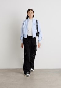 Giacca bomber azzurro chiaro, top bianco e pantaloni cargo neri con tasche. Sneakers nere con strisce bianche. Il modello cammina su uno sfondo neutro.