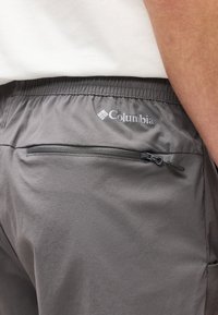 Κοντινή λήψη πίσω τσέπης γκρι παντελονιού Columbia με φερμουάρ και ελαστική μέση, φορεμένο με λευκό πουκάμισο και ορατό χέρι.