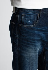 Donkerblauwe denim jeans met een slim fit. Kenmerken zijn een klein geborduurd logo op de achterzak en contrasterende gele stiksels.