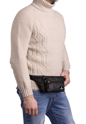 Black nylon waist bag with multiple zippered compartments, worn over blue denim jeans, with a light beige textured sweater.