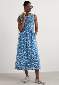Blue patterned midi dress with a smocked bodice, round neckline, and pockets. Model wears light-colored, textured sneakers and oversized sunglasses.