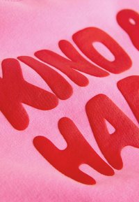 Rosa Stoff mit erhabenen, kräftigen roten Buchstaben in organischer Form. Der Text lautet "KIND OF HARD" und hat insgesamt eine glatte Textur.