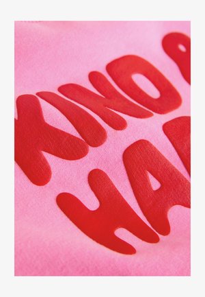 Ružičasta tkanina s izdignutim, podebljanim crvenim slovima u organskom obliku. Tekst glasi "KIND OF HARD" i ima glatku teksturu.