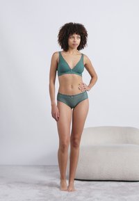 Sutiã verde de renda e calcinhas hipster combinando, com bordas em scallop e um pequeno laço decorativo no centro. Textura lisa, design ajustado.