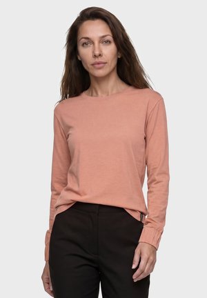 Frau mit langen braunen Haaren, die ein hellrosa Langarmshirt und schwarze Hosen trägt, steht vor einem schlichten hellen Hintergrund.
