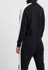 Svart träningsoverall med vita sidosträck. Hög krage, ribbade ärm- och midjeband. Mjuk, medelvikts tyg med avslappnad passform och minimal detaljering.