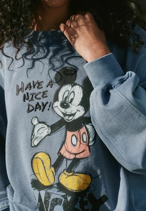 Osoba w niebieskiej bluzie z Myszką Miki i napisem "Miłego dnia!" trzymająca kołnierz przy szyi.