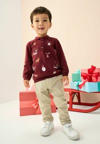 Bordový pletený svetr s vánočním výšivkem, třemi knoflíky a žebrovanými manžetami. K outfitu patří béžové kalhoty a bílé tenisky.