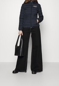 Laivastonsininen toppatakki, jossa vaakasuora tikkaus, edessä vetoketju ja huppu; yhdistetty mustiin leveälahkeisiin housuihin ja teksturoidun mustan käsilaukun kanssa.