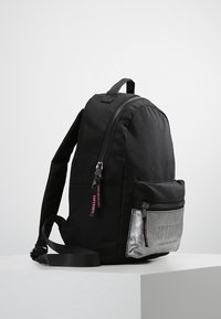 Musta kankainen reppu, jossa on pyöreä muoto, vetoketjullinen pääosasto ja hopeinen etutasku, jossa on kohokuvioitua tekstiä ja vaaleanpunaisia yksityiskohtia.