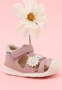 Sandale rose pour enfants avec orteils ouverts, deux décorations florales sur la bride et une marguerite blanche derrière sur fond rose.