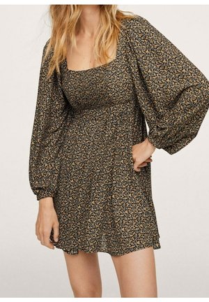 Mujer con un vestido corto de manga larga floral con mangas abullonadas y escote cuadrado, de pie contra un fondo liso.