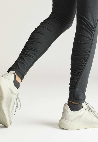 Beine in schwarzen, gerafften Leggings und weißen Sportschuhen gehen über eine weiße Fläche.