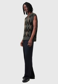 Gilet sans manches tricoté en vert foncé avec un motif en losanges noirs, présentant un col en V et un ourlet côtelé. Porté avec un pantalon et des chaussures noires.