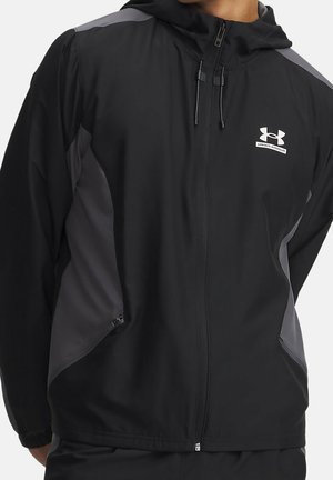 Schwarze und graue Under Armour Kapuzenjacke mit Reißverschluss, Kordelzug und seitlichen Reißverschlusstaschen, getragen von einer Person.