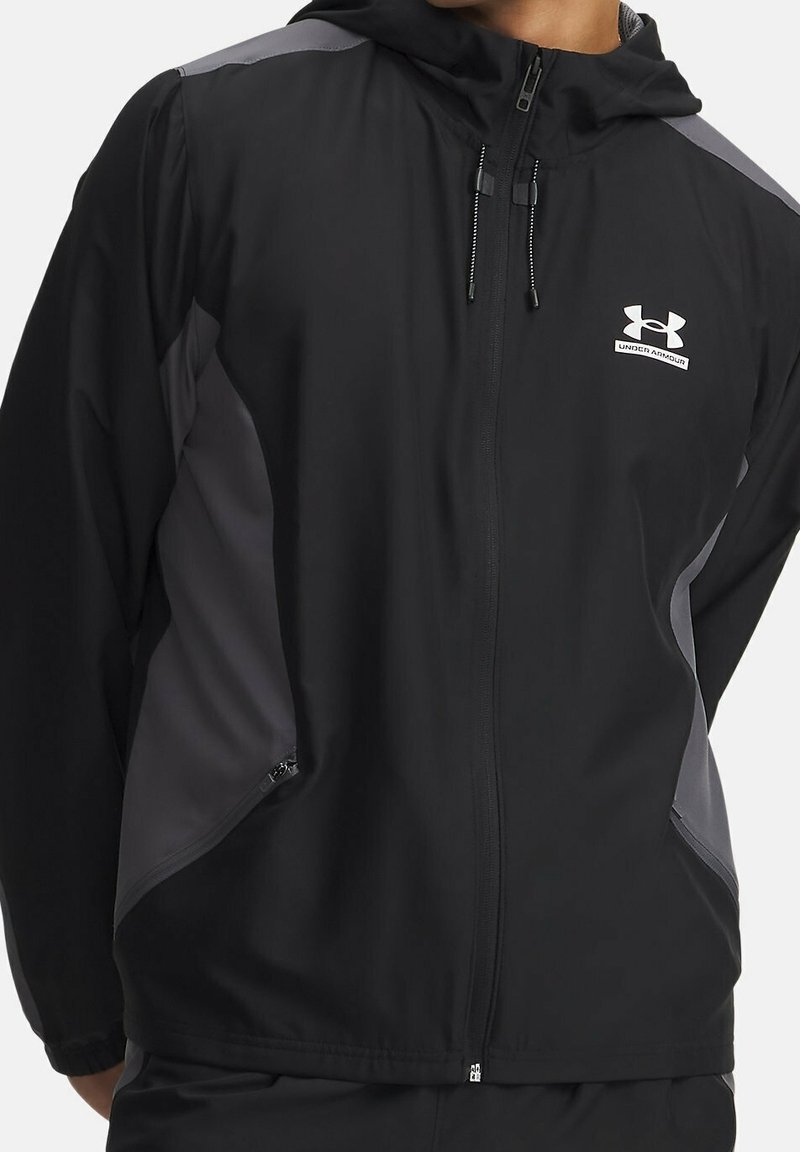 Veste à capuche zippée noire et grise Under Armour avec cordons et poches latérales zippées portée par une personne.