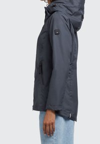 Dunkelblaues Kapuzenjackett aus glattem Stoff, mit einer Reißverschlusstasche an der Seite und einem kleinen Logo-Patch am Ärmel.
