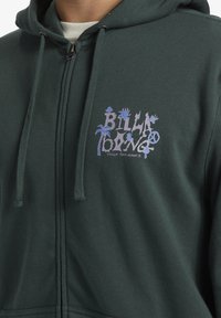 Sudadera con cremallera de color verde oscuro, hecha de una tela texturizada, que presenta un colorido logo gráfico en el lado izquierdo del pecho con elementos caprichosos.