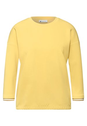 Chemise jaune à manches longues avec col rond, épaules tombantes et poignets à rayures discrètes sur fond blanc.