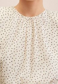 Blouse blanche avec un col plissé, ornée de petits pois noirs sur un tissu léger, créant une texture délicate.