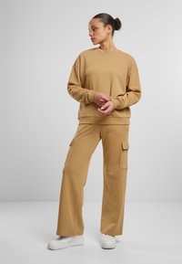 Beige Baumwollpullover mit überschnittenen Schultern, passende Cargo-Hosen mit seitlichen Taschen, ergänzt durch weiße Sneakers.
