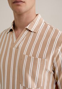 Camisa de manga corta a rayas en tonos beige y blanco, con cuello de pico, tela texturizada y un bolsillo en el pecho.