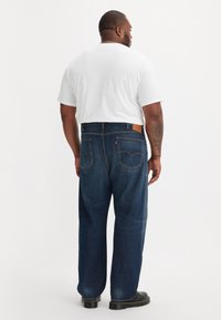 Man in een wit T-shirt met korte mouwen, donkerblauwe jeans en zwarte schoenen, staand met zijn rug naar de camera op een witte achtergrond.