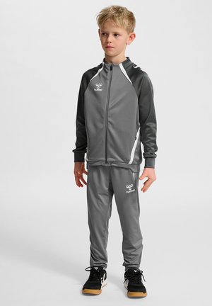 Grauer Sportanzug mit weißen Akzenten; besteht aus einer Reißverschlussjacke und Hose mit elastischem Bund. Getragen mit schwarzen Sneakers.