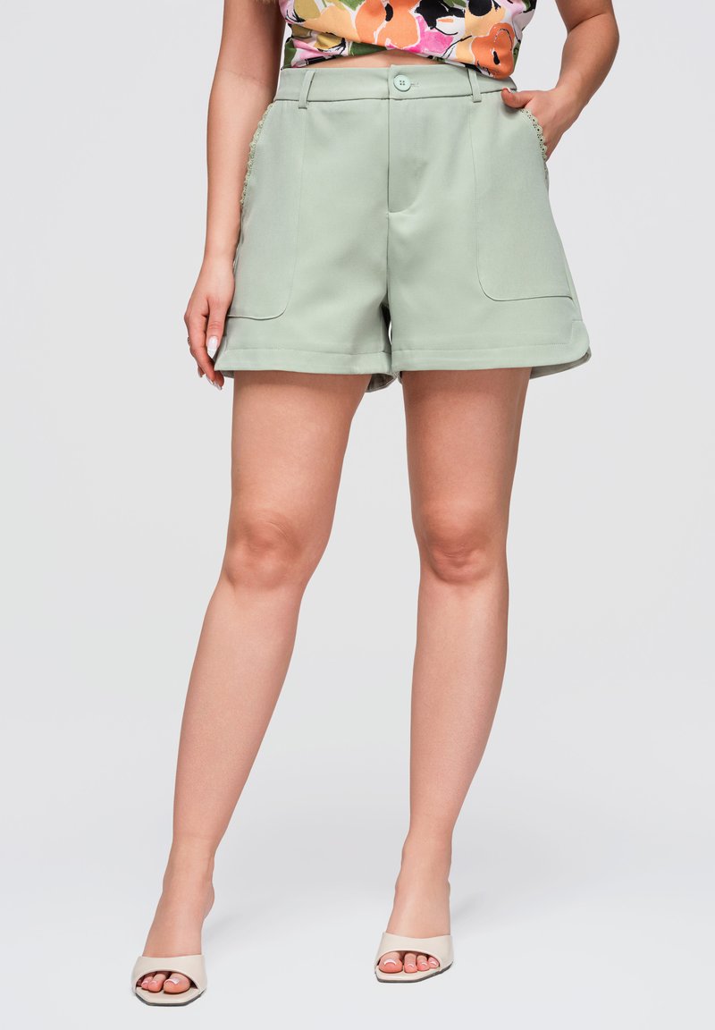Femme portant un short taille haute vert clair avec poches avant et bouton, associé à un crop top fleuri et des talons blancs ouverts aux orteils.