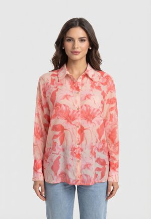 Vrouw draagt een blouse met lange mouwen en knopen, met een roze en koraalkleurige bloemenprint, gecombineerd met lichtblauwe jeans, staand tegen een effen achtergrond.