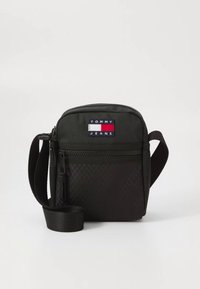 Geantă crossbody din material textil negru cu model texturat, închidere cu fermoar, curea reglabilă și o etichetă cu logo în roșu, alb și albastru.