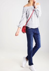 Blusa bianca a spalla scoperta con maniche lunghe e motivo; abbinata a jeans blu e sneaker bianchi; la borsa a tracolla rossa aggiunge un contrasto.