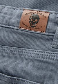 Patch en cuir marron avec un crâne noir et du texte cousu sur un jean en denim bleu clair près de la taille.