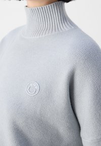 Lichtgrijze gebreide trui met een hoge geribbelde kraag en een geborduurd rond logo op de borst. Zachte textuur met een aangesloten silhouet.