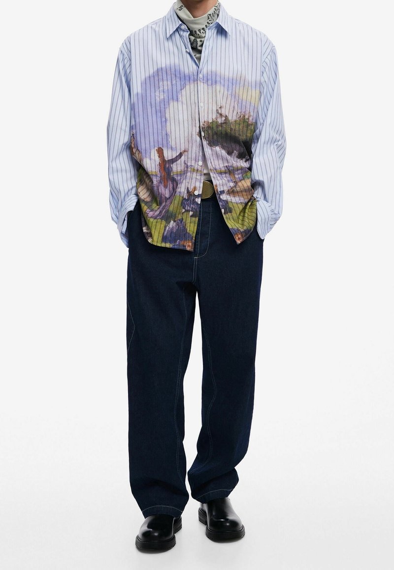 Homme portant une chemise à rayures bleu clair boutonnée avec un imprimé artistique abstrait coloré, un pantalon ample foncé, des chaussures noires et un foulard à motifs.