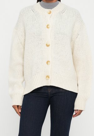 Personne portant un cardigan épais blanc à boutons par-dessus un col roulé gris et un jean bleu foncé, coupé à mi-cuisse.