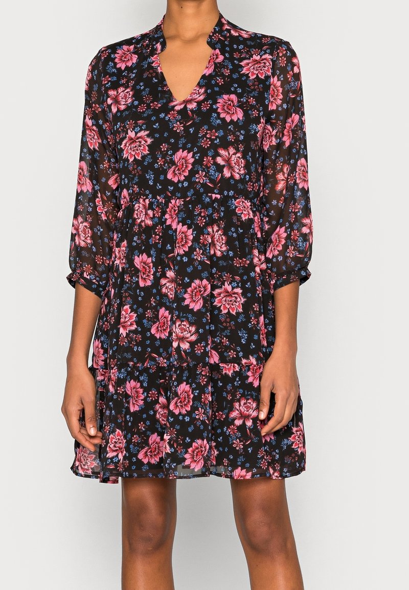 Femme portant une mini robe noire à fleurs avec des fleurs roses et bleues, des manches trois quarts et un col en V.