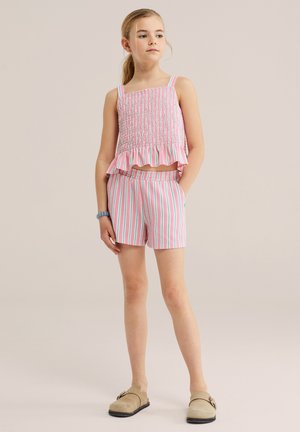 Jong meisje staand, draagt een roze, witte en groene gestreepte mouwloze top met ruchtrand en bijpassende shorts, beige instapschoenen en een armband.