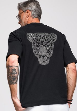Homme aux cheveux gris et lunettes portant un t-shirt noir avec un motif détaillé de visage de léopard argenté dans le dos, montrant son avant-bras droit tatoué.