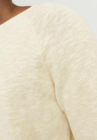 Suéter de punto color beige claro con escote en V, que presenta un patrón texturizado. La tela parece suave y ligeramente translúcida con detalles de costura visibles.