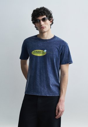 Νέος άνδρας που φοράει σκουρόχρωμα γυαλιά ηλίου και ξεβαμμένο μπλε t-shirt MISBHV με μαύρο παντελόνι, στέκεται μπροστά σε ένα απλό ανοιχτό γκρι φόντο.