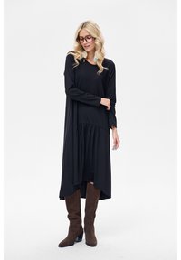 Czarna sukienka z długim rękawem, zaokrąglonym dołem i marszczonym detalem w talii. Modelka ma na sobie brązowe kozaki do kolan i duże okulary. Gładka faktura tkaniny.