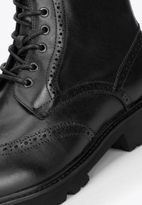 Botín de cuero negro con detalles de brogue, puntera redonda, seis ojales y una suela robusta con surcos pronunciados.