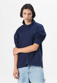 Polo de manga larga azul marino en tejido suave, con cuello abotonado y puños acanalados. Detalle del logo en el pecho, corte relajado.