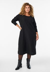 Žena s kovrčavom kosom, odjevena u strukturiranu crnu midi haljinu s dugim rukavima, crne tajice i crne čizme do gležnja, stoji s jednom rukom na bok.