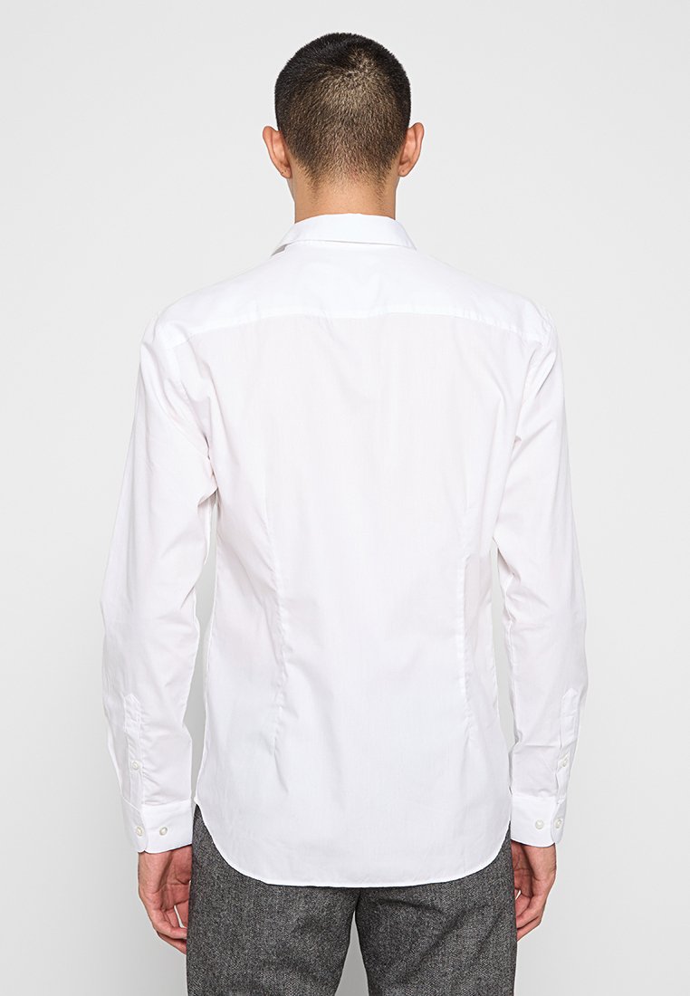 Man gezien van achteren in een nauwsluitend wit langslijfshemd en grijze gestructureerde broek tegen een effen lichte achtergrond.