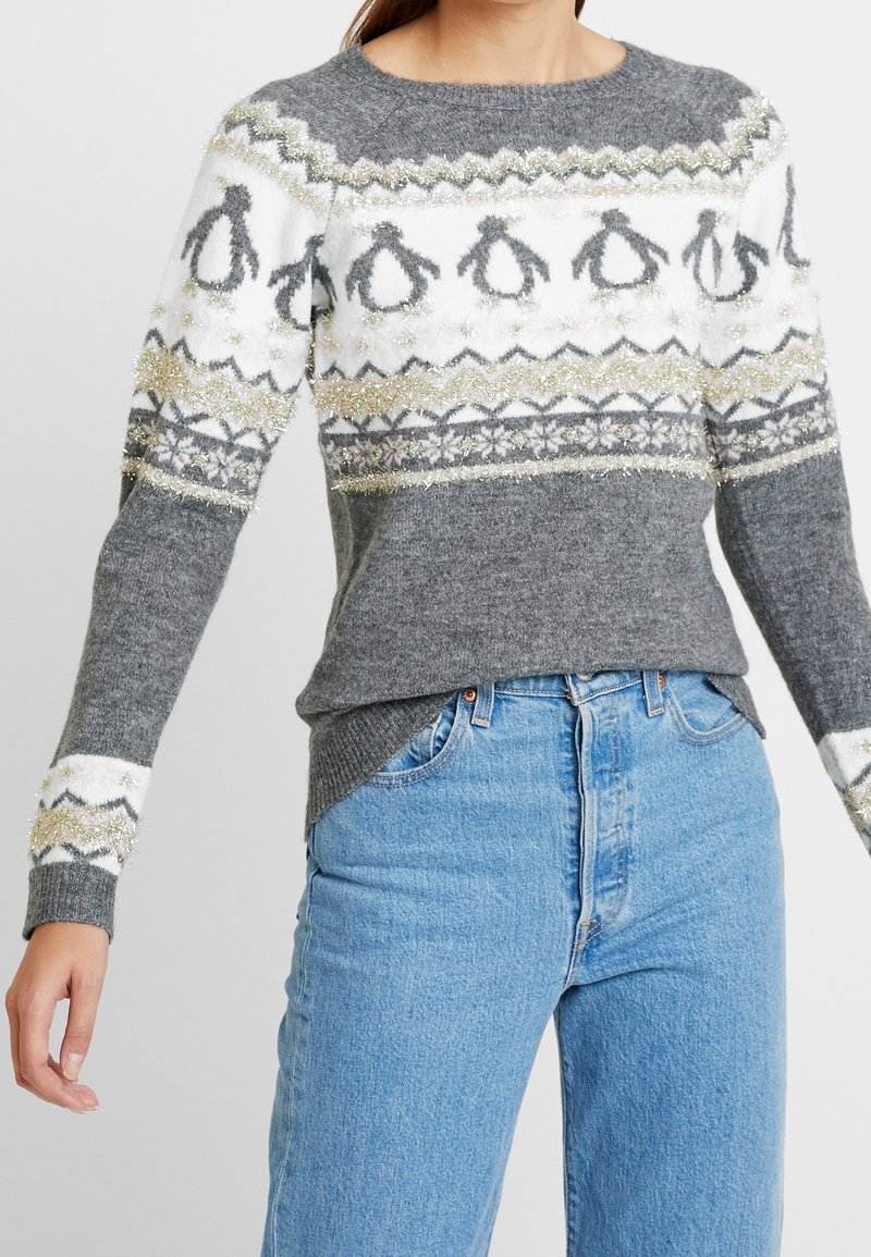 Femme portant un pull en maille gris et blanc avec des motifs de pingouins et de flocons de neige, associé à un jean bleu clair.
