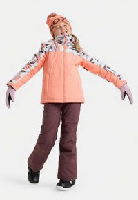 Kind lächelt mit ausgestreckten Armen, trägt eine rosa Winterjacke mit Muster, bordeauxrote Schneehose, orangefarbene Mütze, violette Handschuhe und schwarze Stiefel.