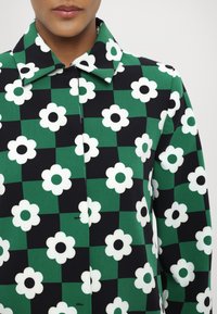 Cappotto con stampa floreale caratterizzato da un motivo a scacchi nero e verde con fiori bianchi, centro nero e un classico design con colletto.