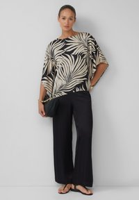 Losse, korte mouwen blouse met een beige en zwart palmbladpatroon; gecombineerd met zwarte wijde broek en zwarte slippers.